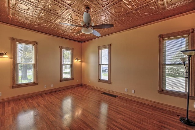 empty room featuring plenty of natural light, a ceiling fan, visible vents, wood finished floors, and an ornate ceiling