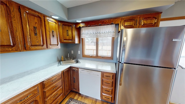 kitchen featuring white dishwasher, a sink, light countertops, freestanding refrigerator, and brown cabinetry