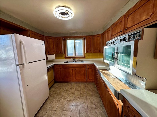 kitchen featuring a sink, light countertops, freestanding refrigerator, brown cabinets, and paneled dishwasher