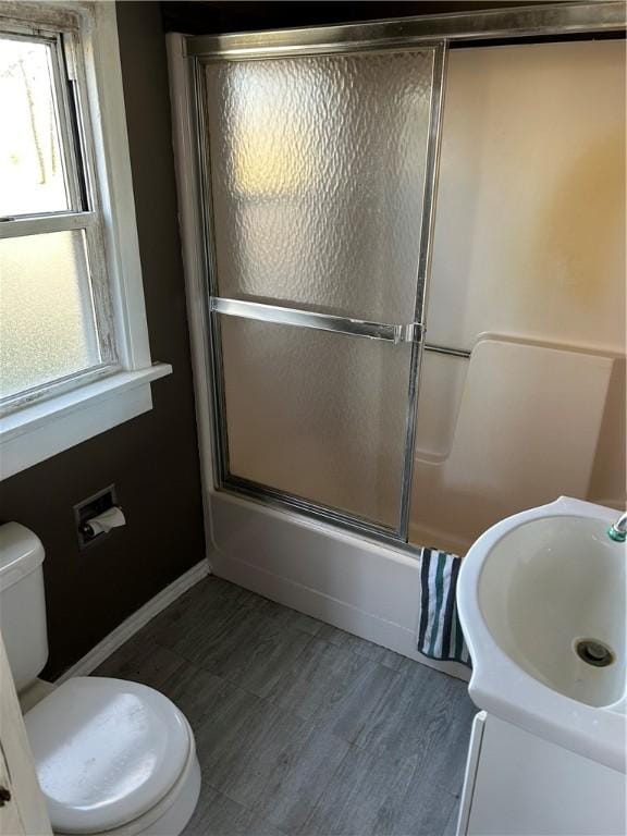 bathroom with toilet, a sink, wood finished floors, bath / shower combo with glass door, and baseboards