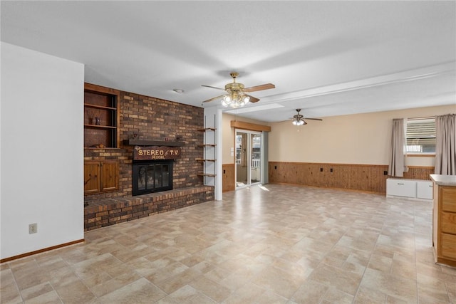 unfurnished living room featuring wooden walls, built in features, a wainscoted wall, ceiling fan, and a brick fireplace