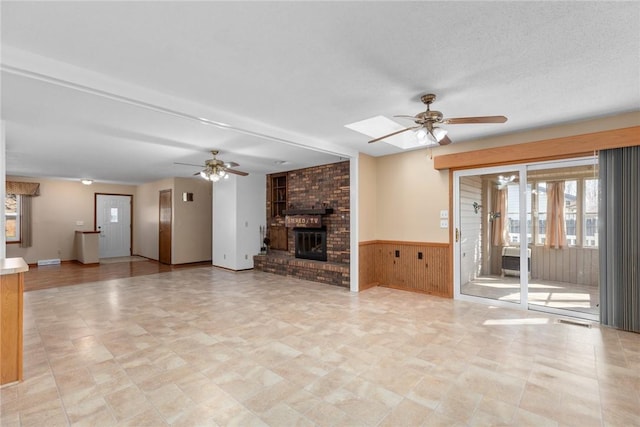 unfurnished living room with a brick fireplace, ceiling fan, a wainscoted wall, wood walls, and a skylight