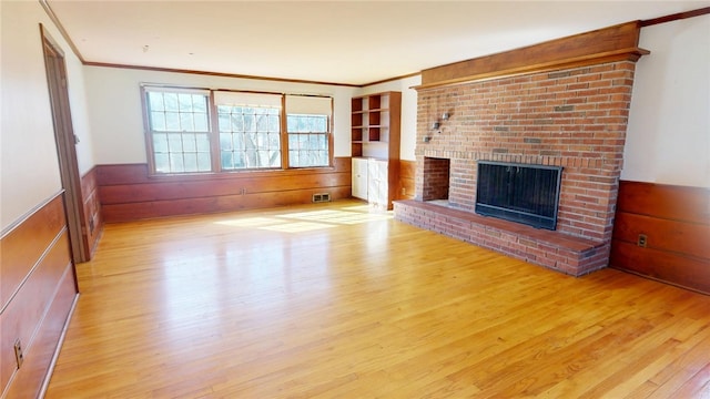 unfurnished living room with a wainscoted wall, wood walls, wood finished floors, a brick fireplace, and crown molding