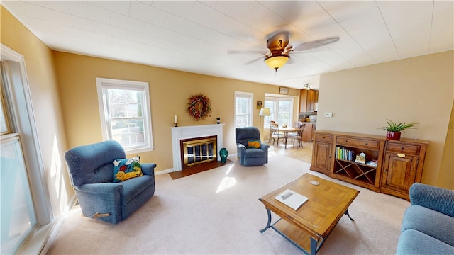 living area with a fireplace with flush hearth, a ceiling fan, light colored carpet, and baseboards