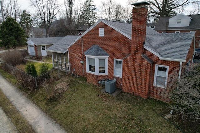 rear view of property featuring roof with shingles, a sunroom, brick siding, central AC unit, and a chimney