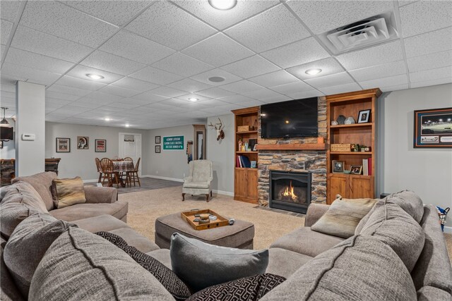 carpeted living room with a drop ceiling and a stone fireplace