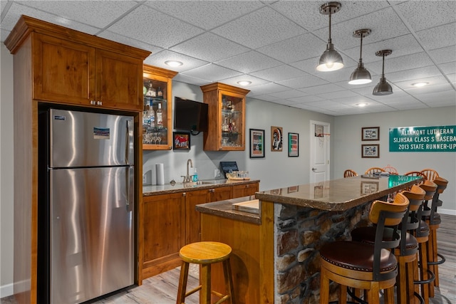 bar featuring sink, stainless steel fridge, dark stone countertops, decorative light fixtures, and light hardwood / wood-style floors