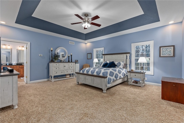 bedroom with a raised ceiling, connected bathroom, ceiling fan, and light colored carpet