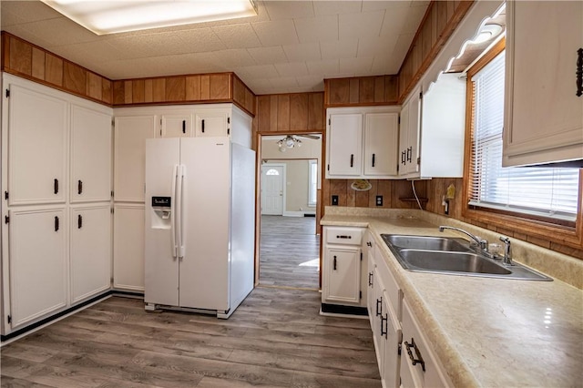 kitchen featuring wooden walls, white fridge with ice dispenser, wood finished floors, white cabinetry, and a sink