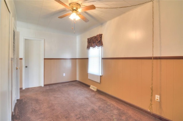 carpeted empty room with visible vents, a ceiling fan, and a wainscoted wall