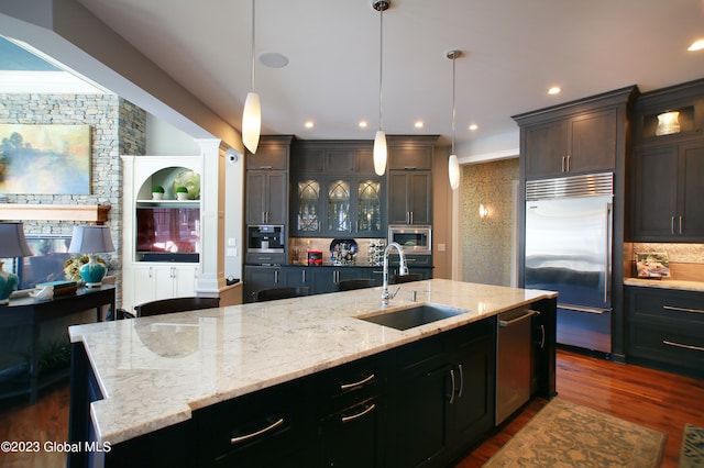 kitchen with built in appliances, decorative light fixtures, dark wood-type flooring, a kitchen island with sink, and sink