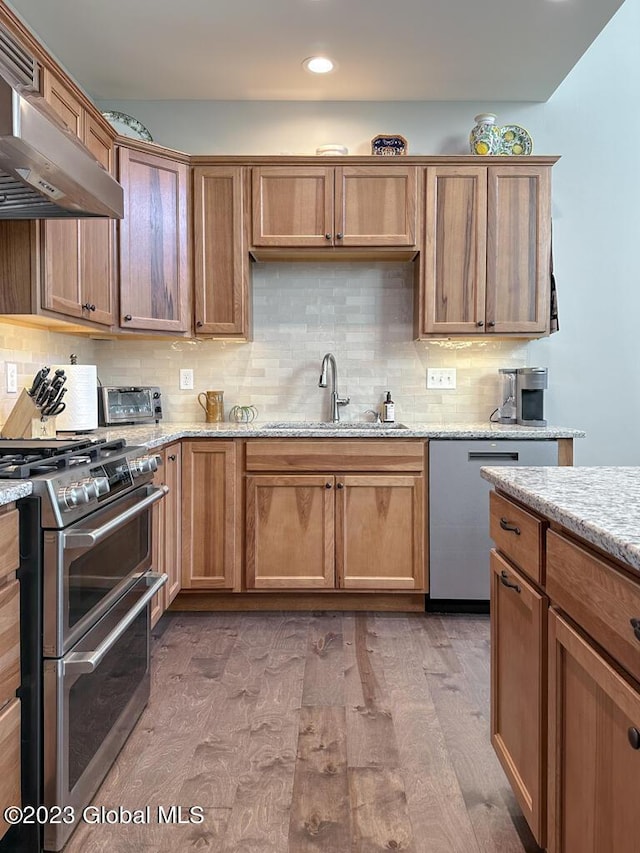 kitchen with wood finished floors, a sink, stainless steel appliances, under cabinet range hood, and backsplash