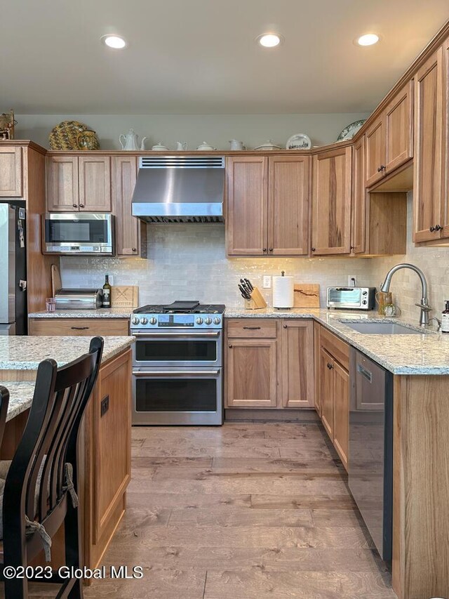 kitchen featuring stainless steel appliances, a sink, light wood-style floors, wall chimney range hood, and light stone countertops