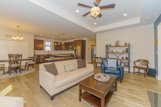 living room with baseboards, light wood finished floors, a tray ceiling, recessed lighting, and ceiling fan with notable chandelier