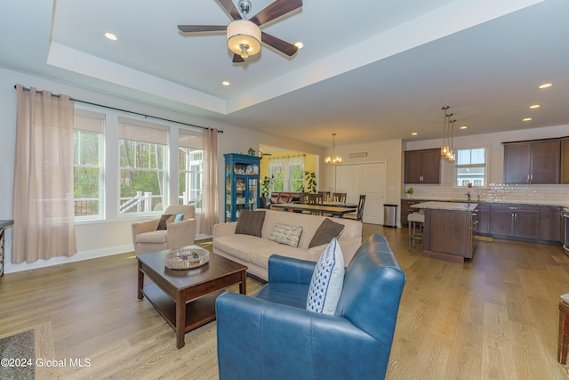 living room with light wood finished floors, baseboards, recessed lighting, ceiling fan with notable chandelier, and a raised ceiling