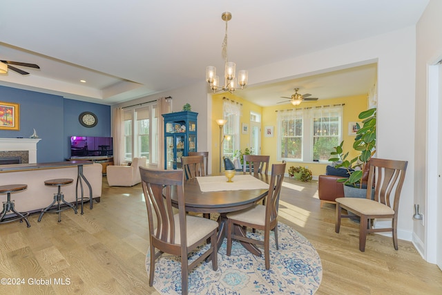 dining area featuring baseboards, a tray ceiling, ceiling fan with notable chandelier, a fireplace, and light wood-style floors