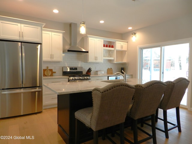 kitchen with decorative light fixtures, light hardwood / wood-style flooring, an island with sink, stainless steel appliances, and wall chimney exhaust hood