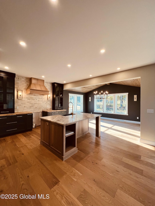 kitchen featuring light hardwood / wood-style flooring, backsplash, light stone counters, custom range hood, and a center island with sink