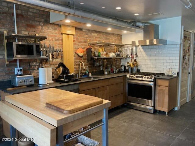 kitchen with dark tile patterned floors, wall chimney range hood, brick wall, stainless steel appliances, and sink