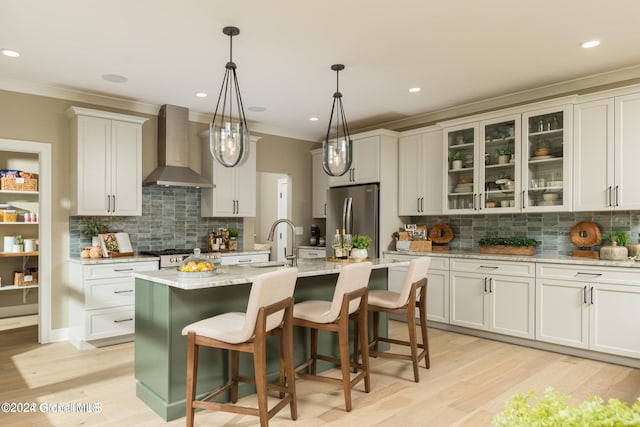 kitchen featuring a kitchen island with sink, wall chimney range hood, decorative backsplash, appliances with stainless steel finishes, and light wood-type flooring