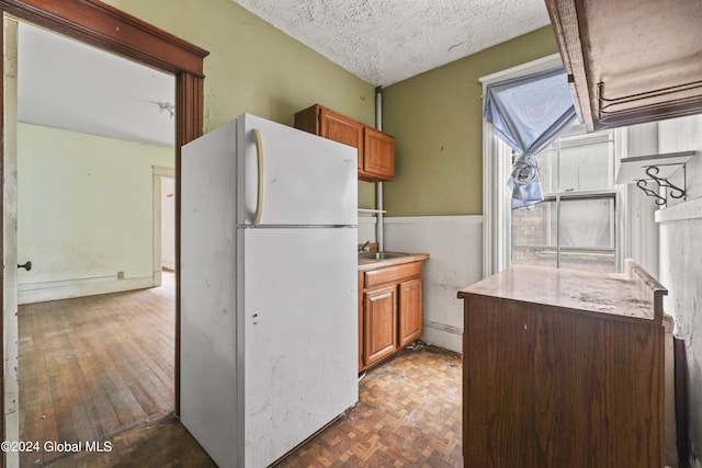 kitchen with parquet flooring, brown cabinetry, a textured ceiling, and freestanding refrigerator