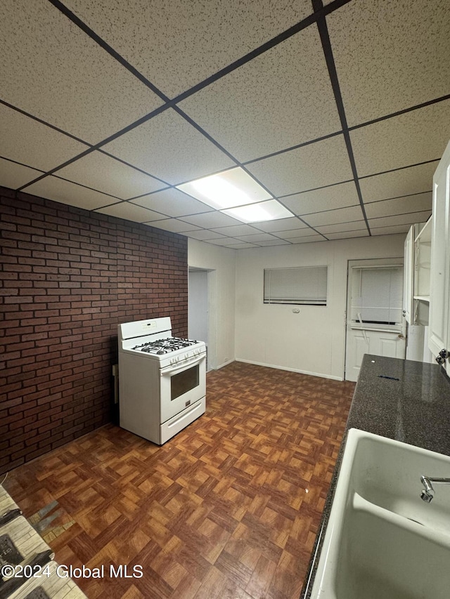 kitchen with brick wall, white gas range oven, white cabinetry, dark parquet floors, and a drop ceiling