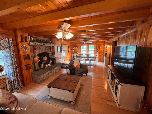 living room with light hardwood / wood-style floors, a stone fireplace, wood walls, beamed ceiling, and ceiling fan