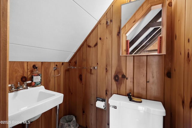 bathroom with vaulted ceiling, wooden walls, toilet, and a sink