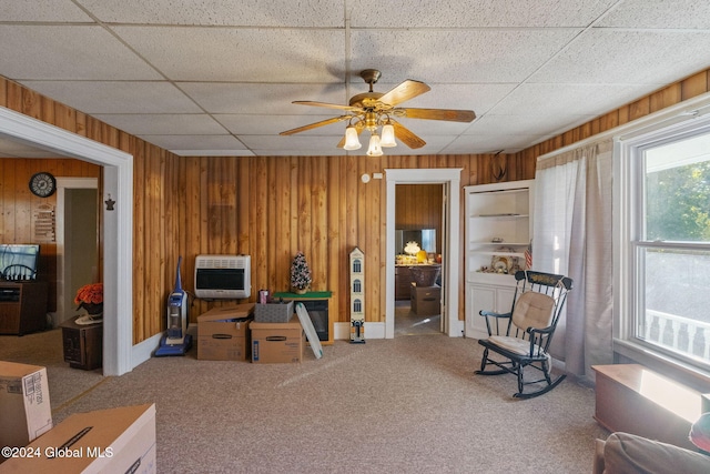 living area featuring ceiling fan, wood walls, heating unit, a paneled ceiling, and carpet