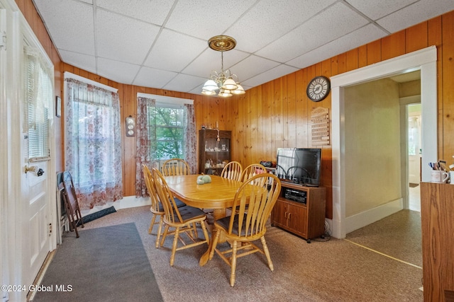 carpeted dining space with a paneled ceiling, wood walls, and an inviting chandelier