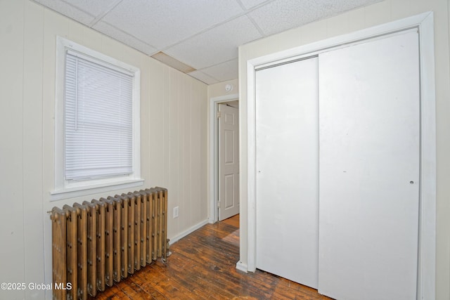 unfurnished bedroom with a paneled ceiling, dark wood finished floors, a closet, radiator heating unit, and baseboards
