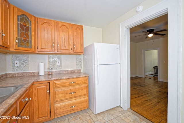 kitchen with glass insert cabinets, freestanding refrigerator, a ceiling fan, brown cabinetry, and decorative backsplash