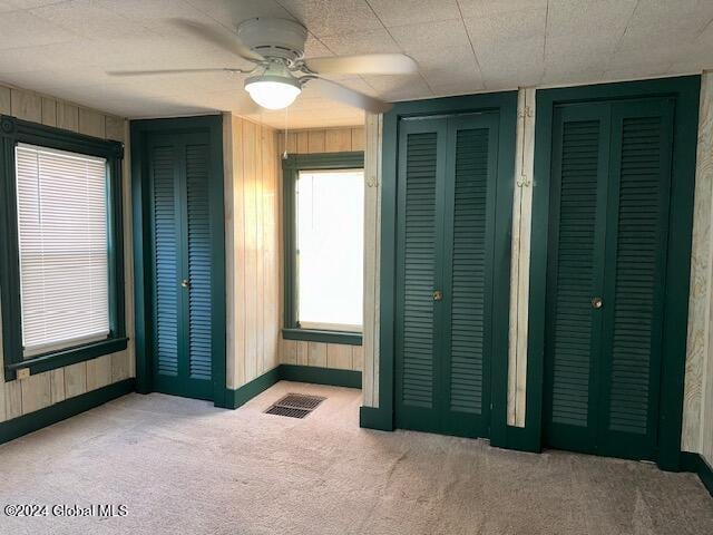 unfurnished bedroom featuring multiple closets, ceiling fan, light colored carpet, and wood walls