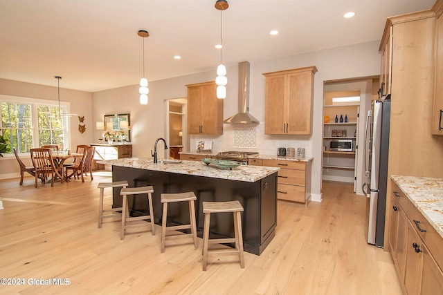 kitchen featuring light stone counters, stainless steel appliances, wall chimney range hood, pendant lighting, and a center island with sink