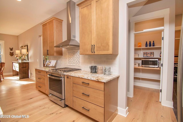 kitchen featuring wall chimney exhaust hood, stainless steel gas range oven, light stone counters, backsplash, and light brown cabinetry