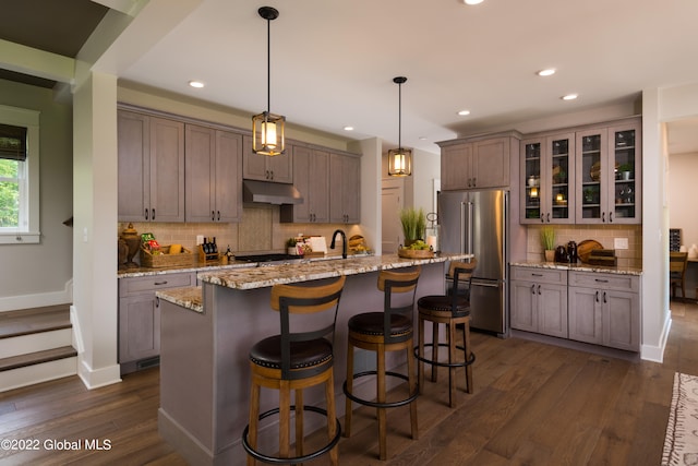 kitchen featuring a center island with sink, dark hardwood / wood-style floors, light stone countertops, high quality fridge, and decorative light fixtures