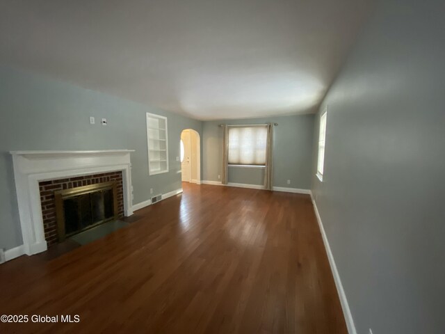 unfurnished living room featuring arched walkways, wood finished floors, visible vents, baseboards, and a brick fireplace