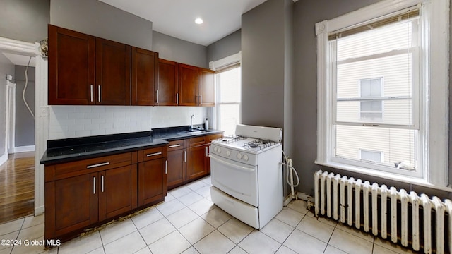 kitchen with backsplash, white gas range, sink, light tile patterned floors, and radiator heating unit