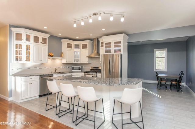 kitchen with wall chimney exhaust hood, light wood-type flooring, a kitchen island, white cabinetry, and stainless steel appliances