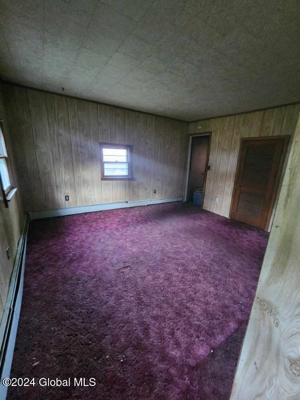 spare room with baseboard heating, dark colored carpet, and wooden walls