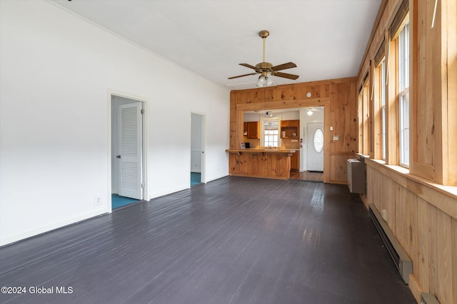 unfurnished living room with dark hardwood / wood-style floors, a baseboard radiator, ceiling fan, and wooden walls