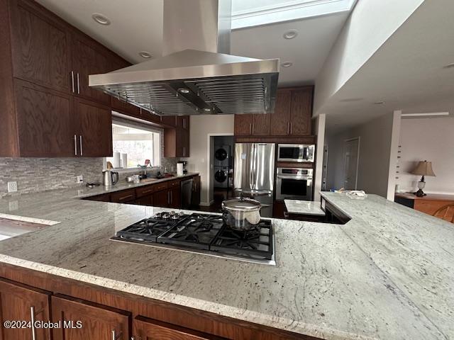 kitchen featuring island exhaust hood, decorative backsplash, light stone countertops, stainless steel appliances, and sink