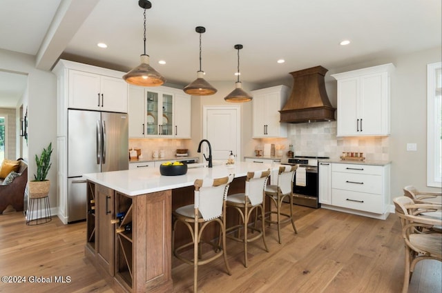 kitchen with white cabinetry, light hardwood / wood-style flooring, a center island with sink, custom exhaust hood, and appliances with stainless steel finishes