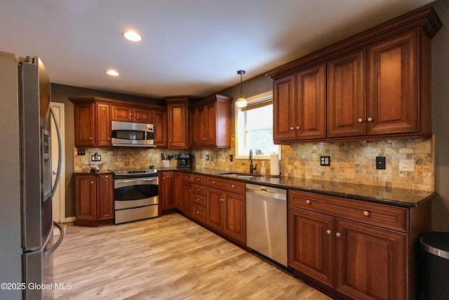 kitchen with stainless steel appliances, sink, light hardwood / wood-style flooring, dark stone countertops, and hanging light fixtures