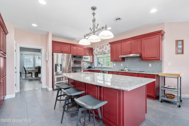 kitchen with a center island with sink, sink, appliances with stainless steel finishes, decorative light fixtures, and light stone counters