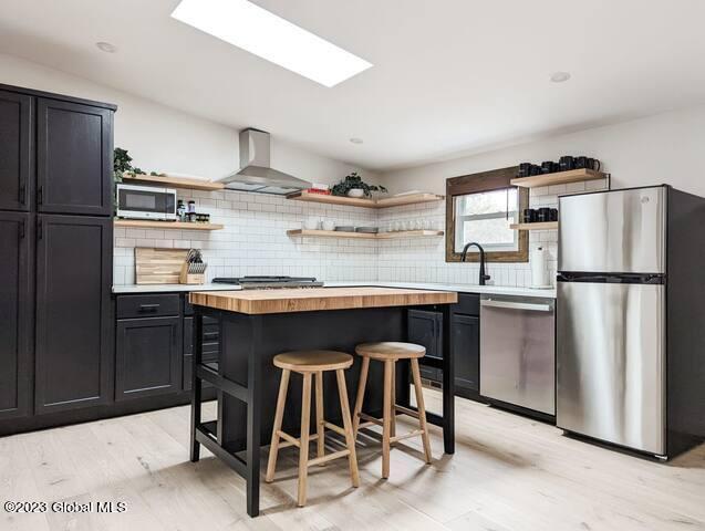 kitchen featuring wood counters, a kitchen breakfast bar, decorative backsplash, stainless steel appliances, and wall chimney range hood