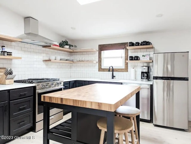 kitchen featuring butcher block counters, wall chimney range hood, a kitchen breakfast bar, backsplash, and appliances with stainless steel finishes