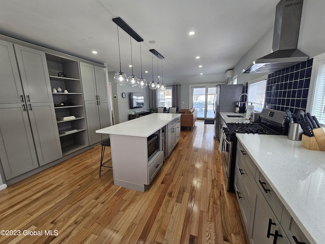 kitchen with hanging light fixtures, wall chimney exhaust hood, gray cabinets, light wood-type flooring, and stainless steel appliances