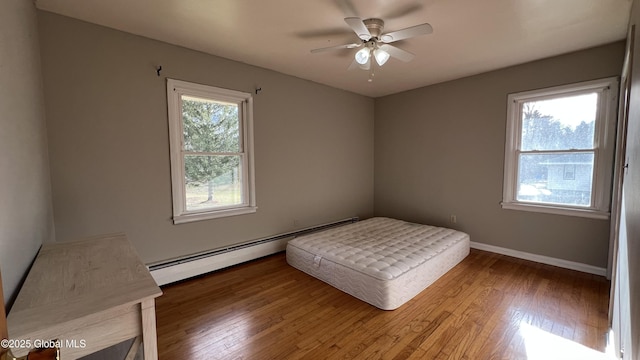 unfurnished bedroom featuring multiple windows, wood-type flooring, ceiling fan, and a baseboard heating unit