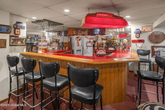 bar with a paneled ceiling, carpet flooring, and white fridge with ice dispenser